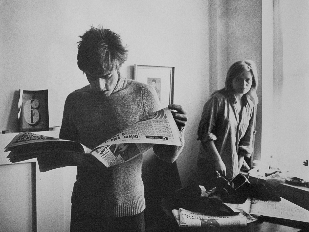 A man and a woman standing in a room looking at a newspaper.