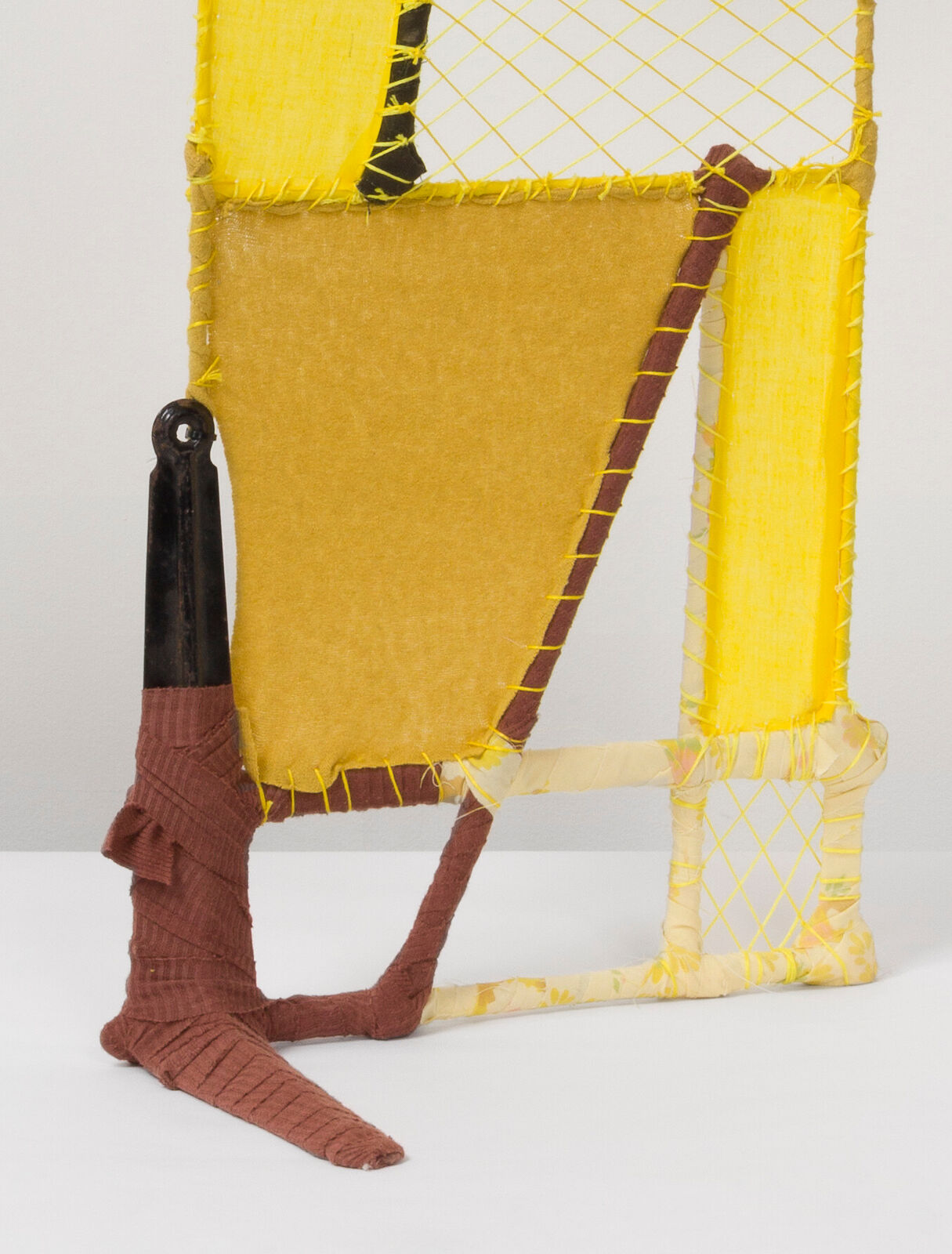 A geometric textile sculpture resembling a stylized house or sailboat, constructed from yellow and brown yarns.