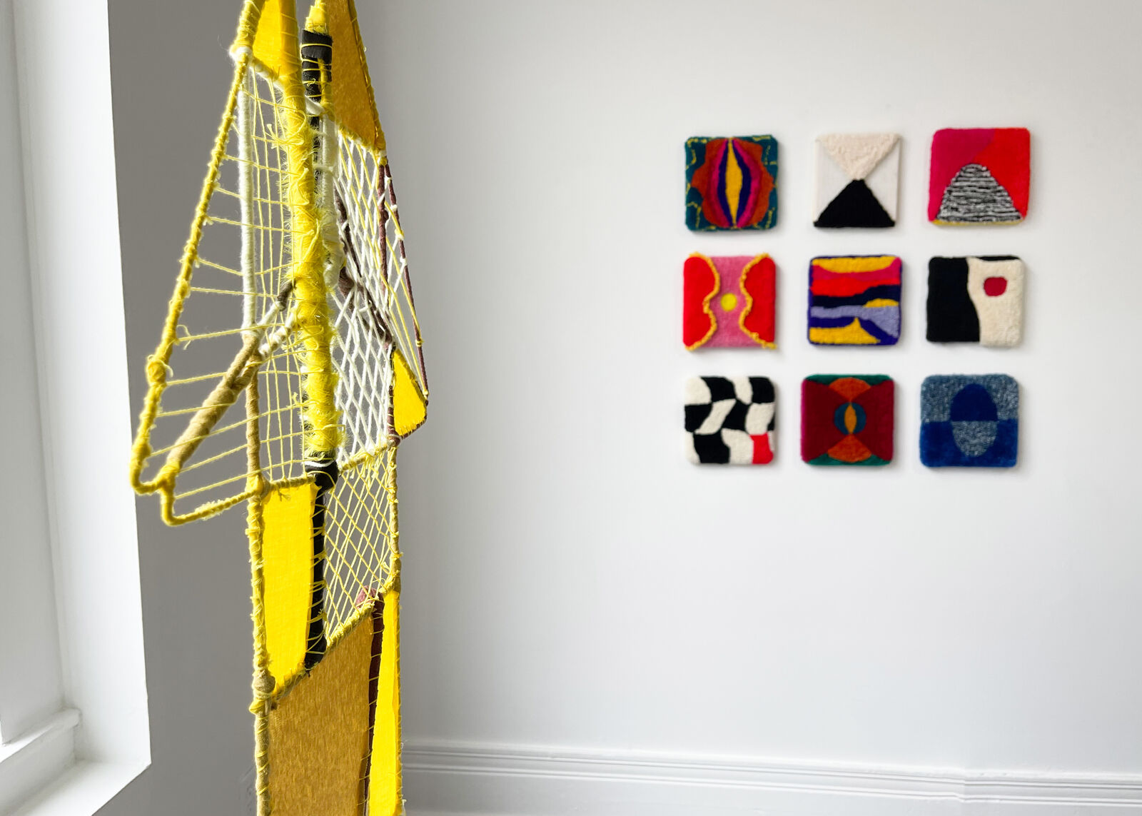 A geometric textile sculpture resembling a stylized house or sailboat, constructed from yellow and brown yarns.