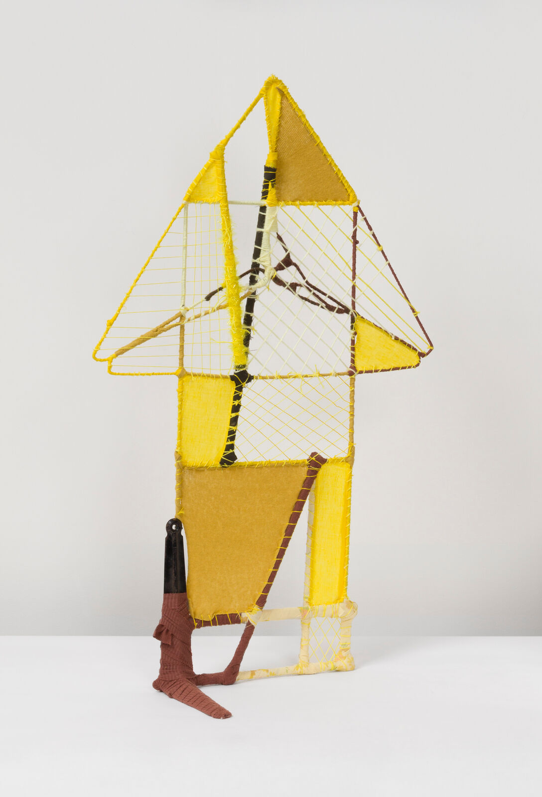 A geometric textile sculpture resembling a stylized house or sailboat, constructed from yellow and brown yarns.