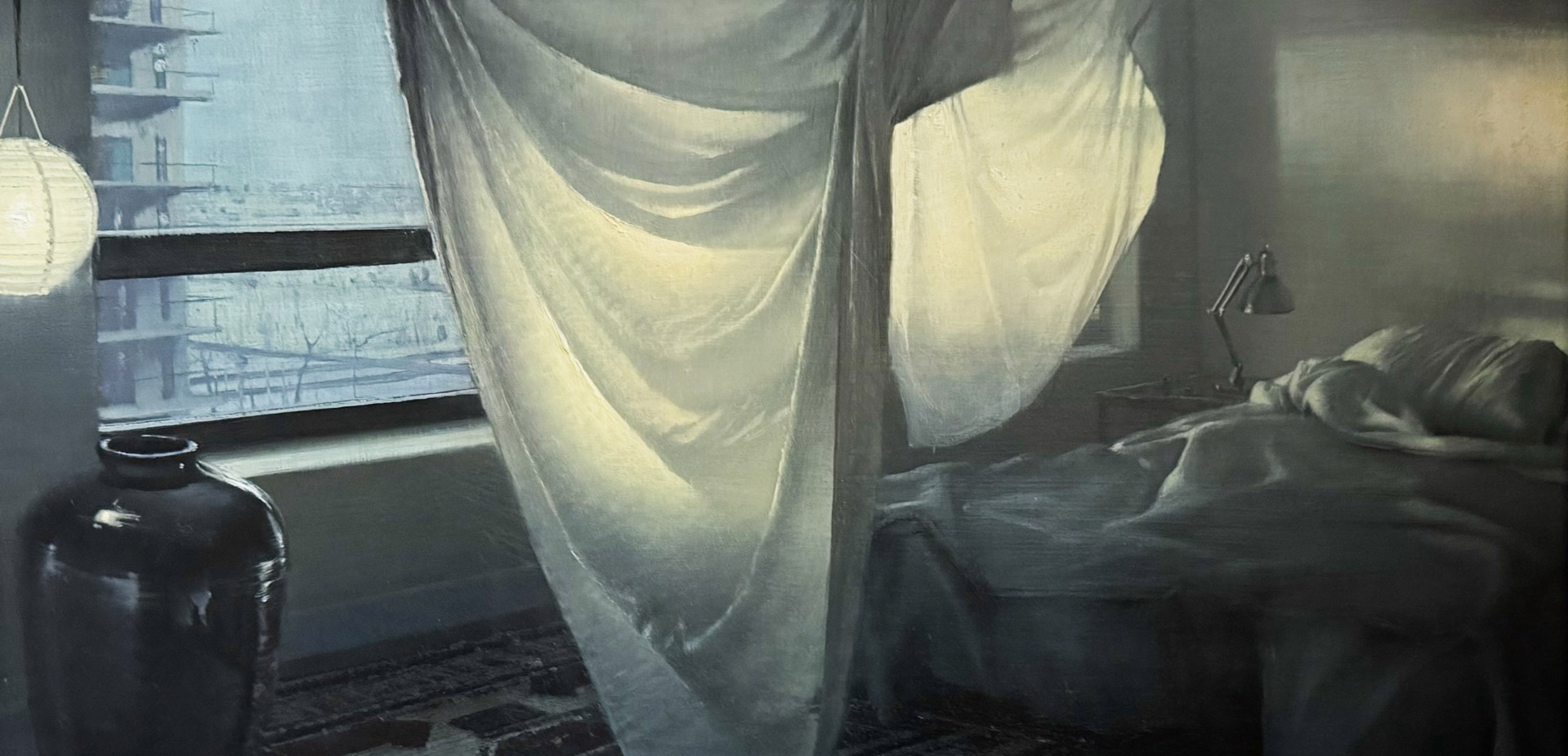 A moody interior scene with a bed, sheer curtains, and a window overlooking an urban landscape.
