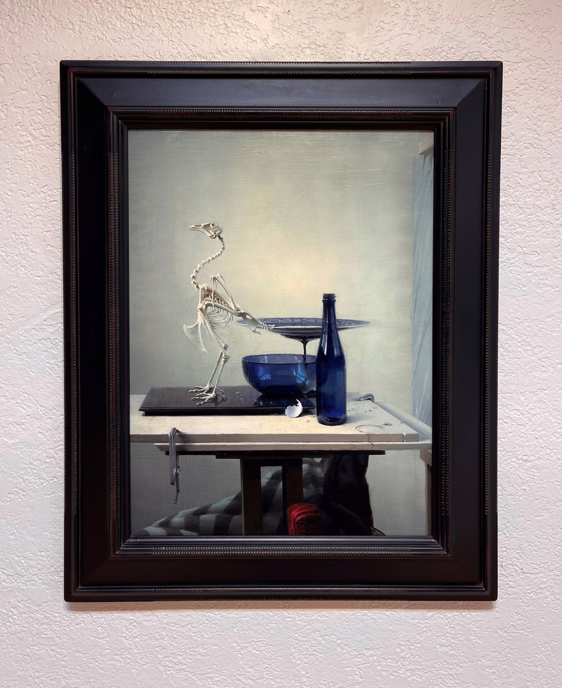 A surreal still life featuring a bird skeleton, blue glassware, and a bottle on a table.