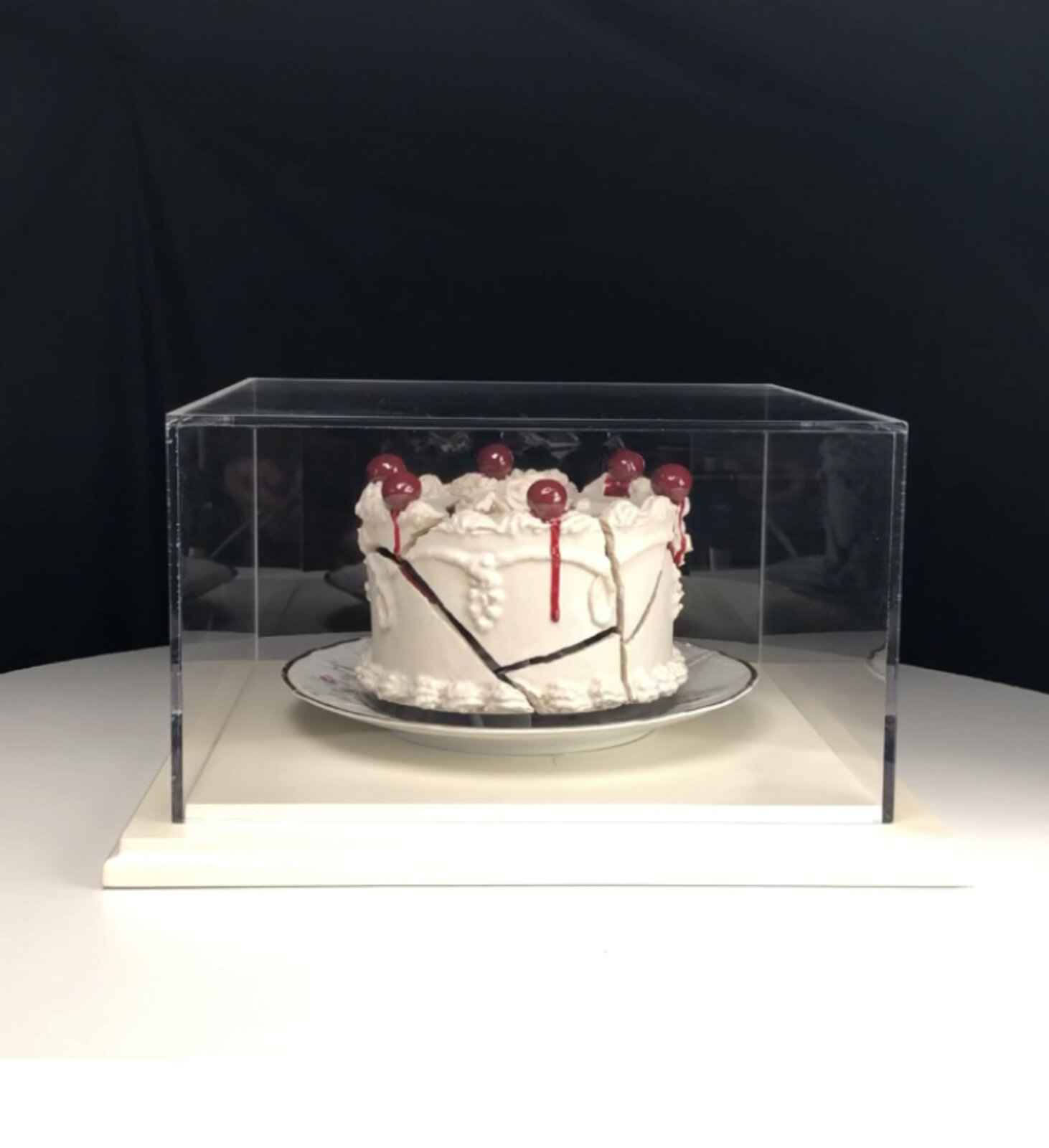 A white frosted cake with red cherries and dark cracks sits inside a clear display case against a black background.
