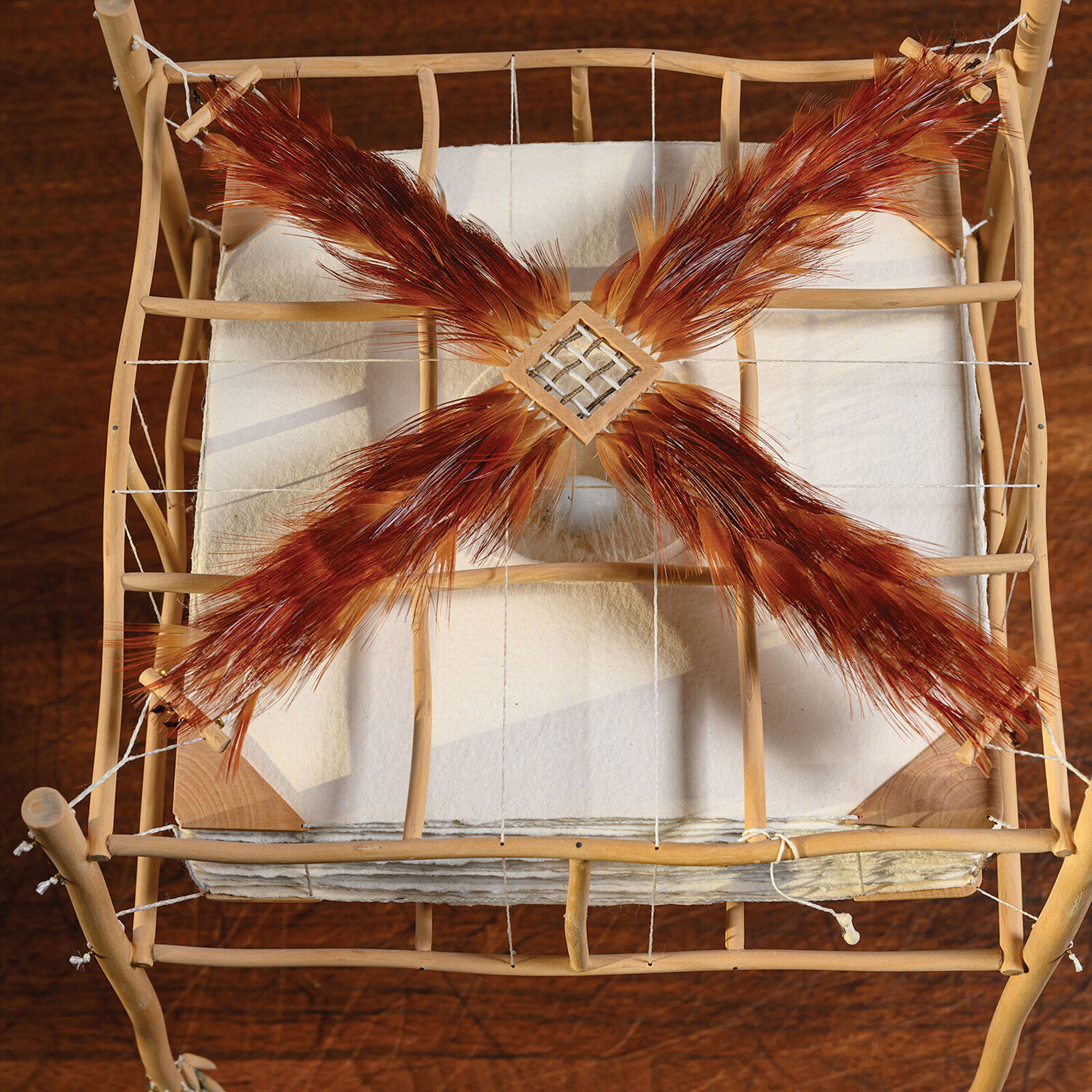 A sculptural installation featuring a woven basket suspended from a bamboo frame with feathered elements.