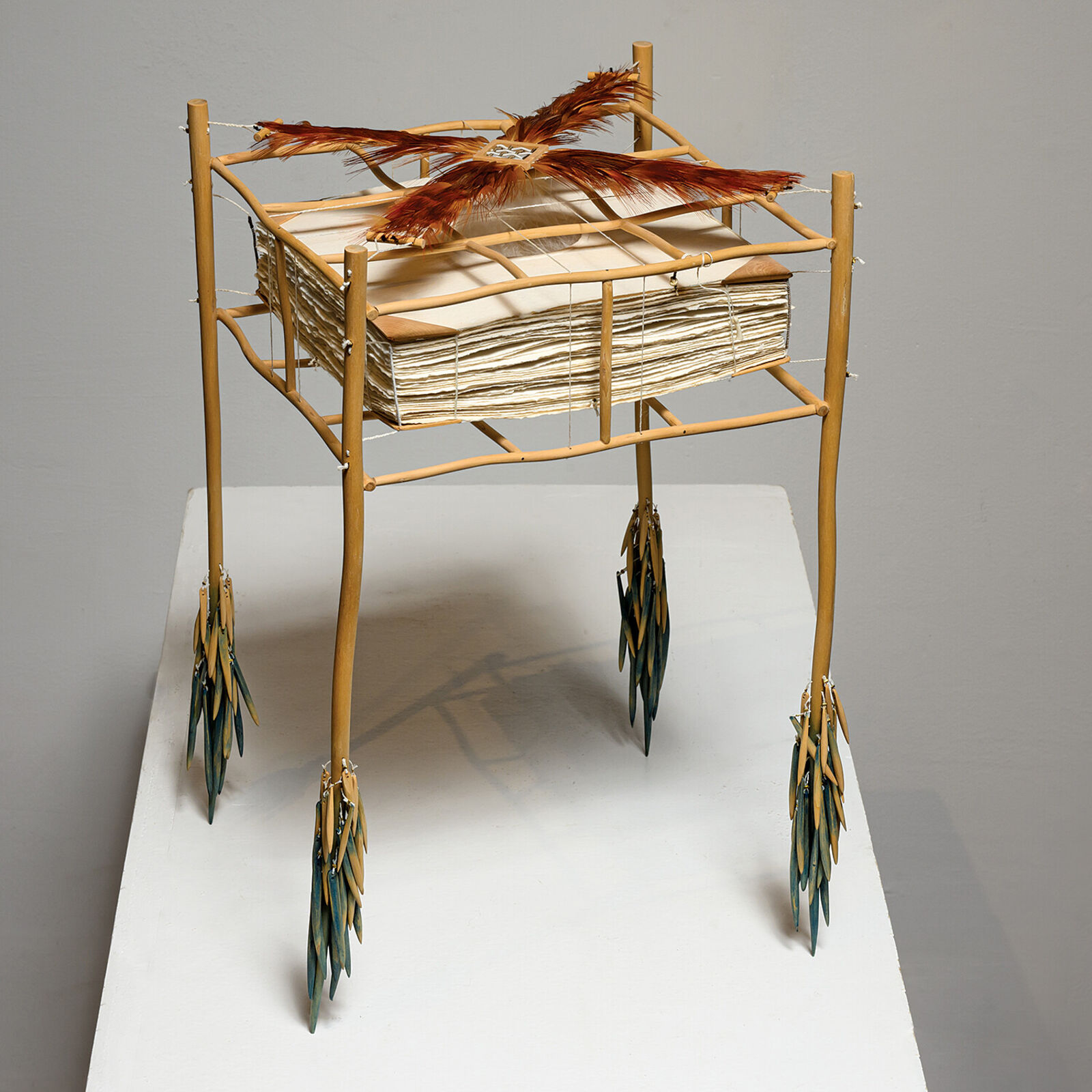 A sculptural installation featuring a woven basket suspended from a bamboo frame with feathered elements.