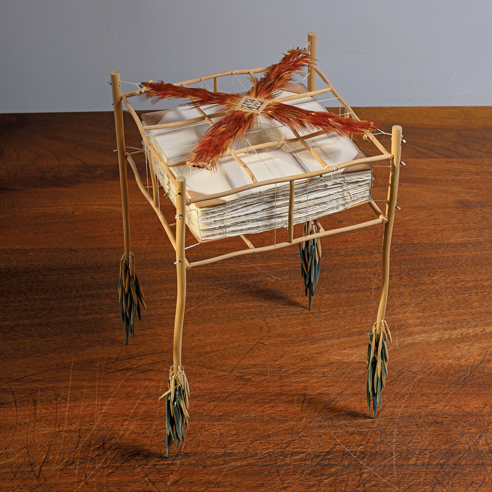 A sculptural installation featuring a woven basket suspended from a bamboo frame with feathered elements.