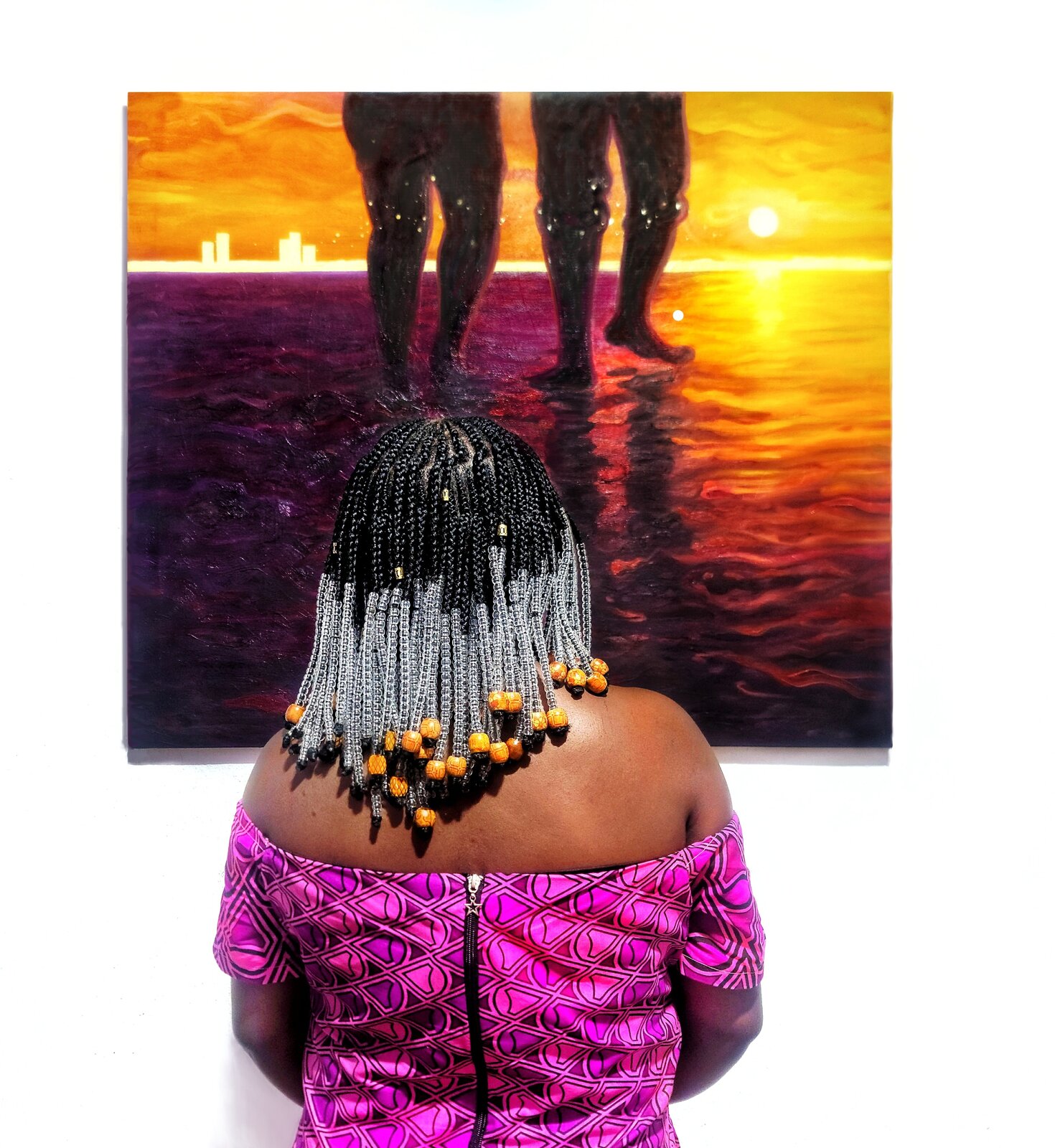 "A pair of figures stand barefoot in the water at sunset, their silhouettes reflected in the rippling surface against a vibrant orange sky and distant city skyline."