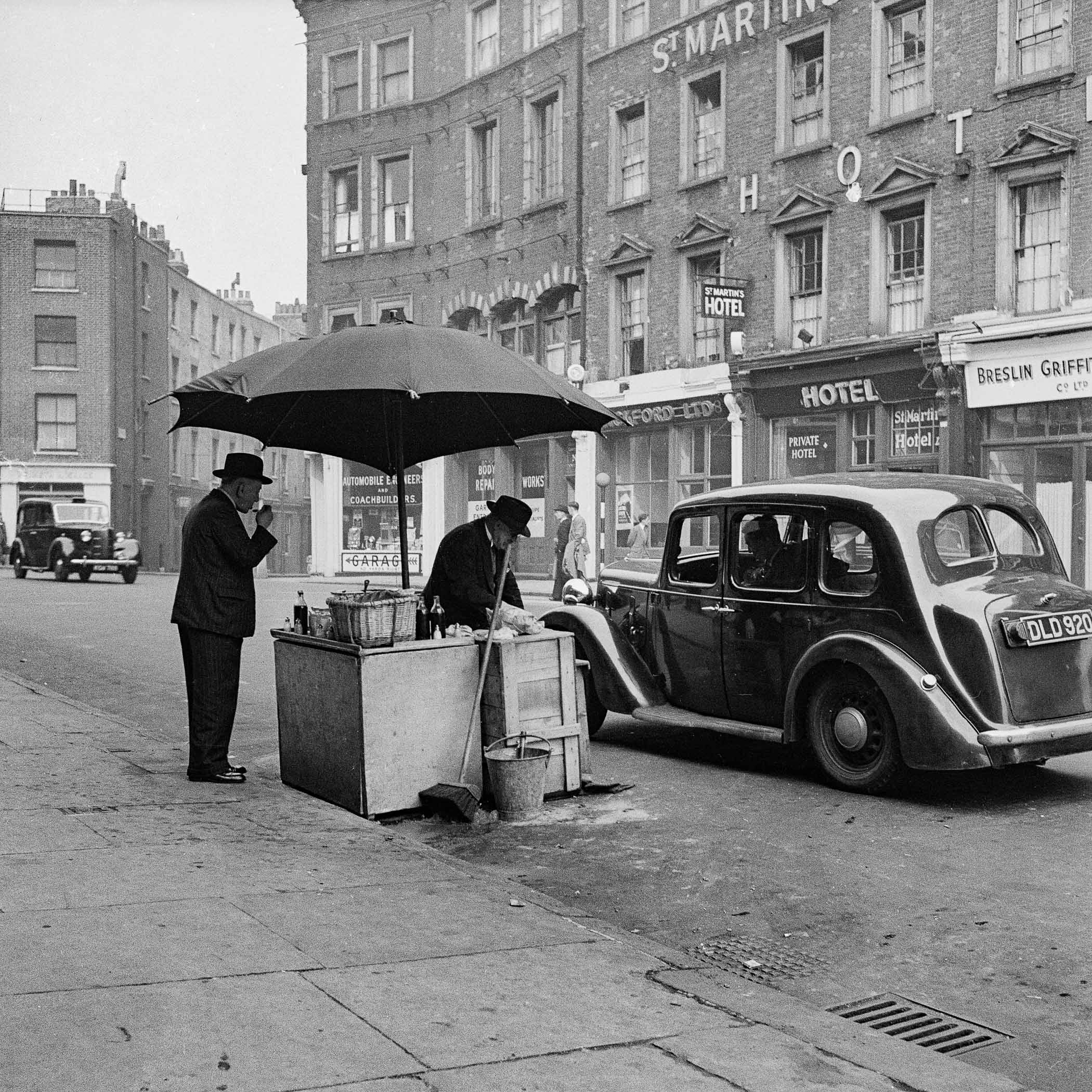 A black and white photo of two men standing under an umbrella on a city street.