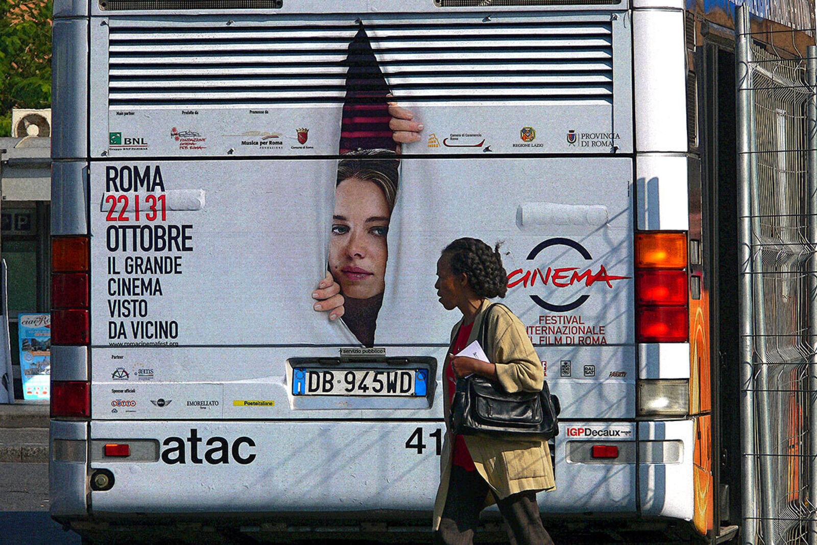 A woman walking past a bus with a poster on the back.