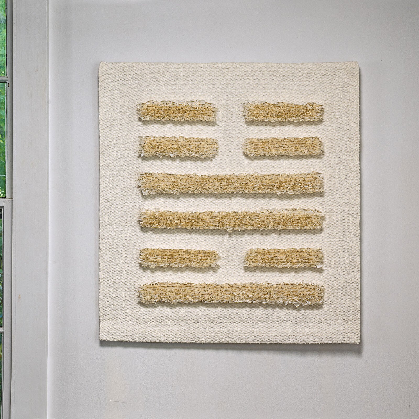 A minimalist textile piece featuring a woven white background with horizontal strips of textured, golden-brown fibers.