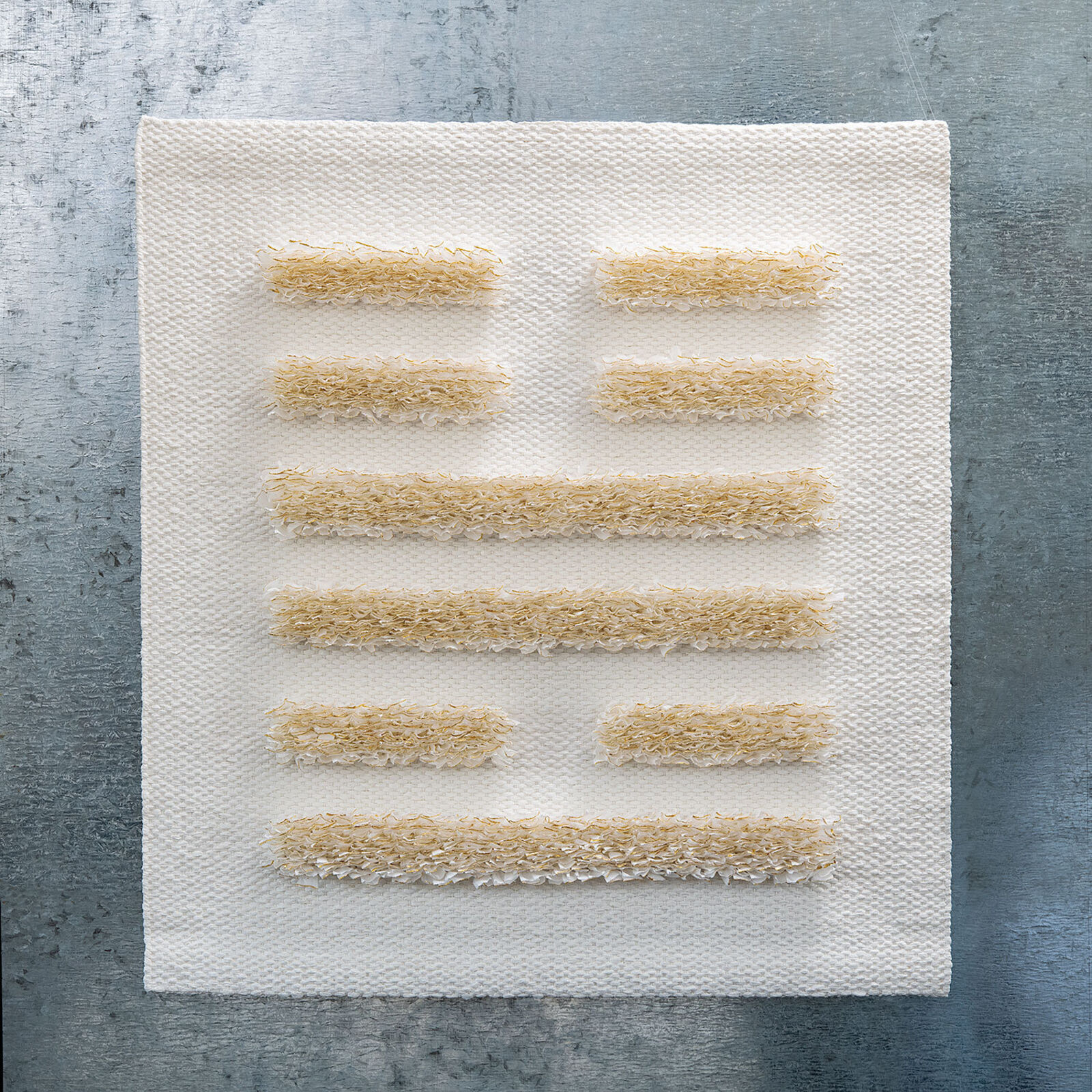 A minimalist textile piece featuring a woven white background with horizontal strips of textured, golden-brown fibers.