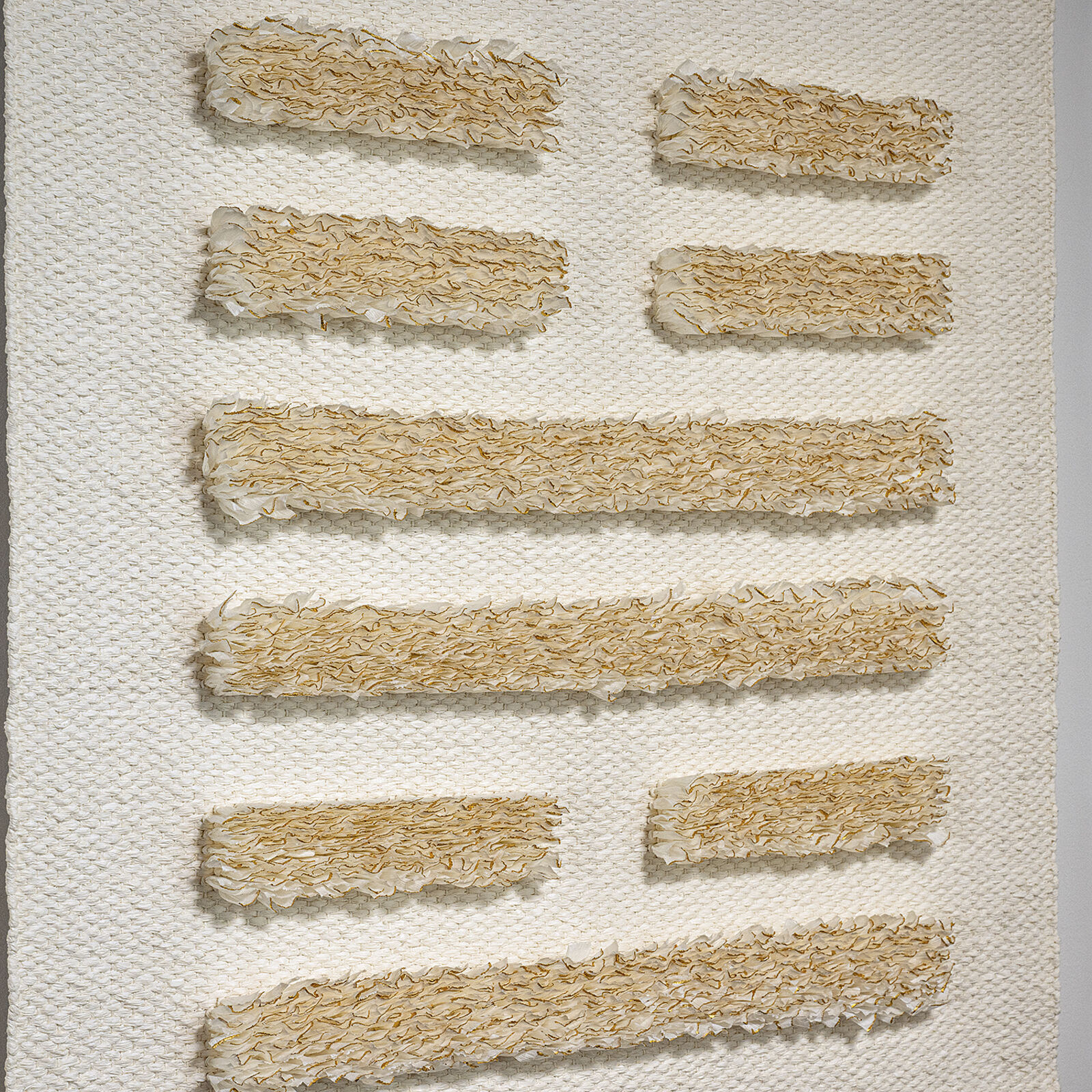 A minimalist textile piece featuring a woven white background with horizontal strips of textured, golden-brown fibers.