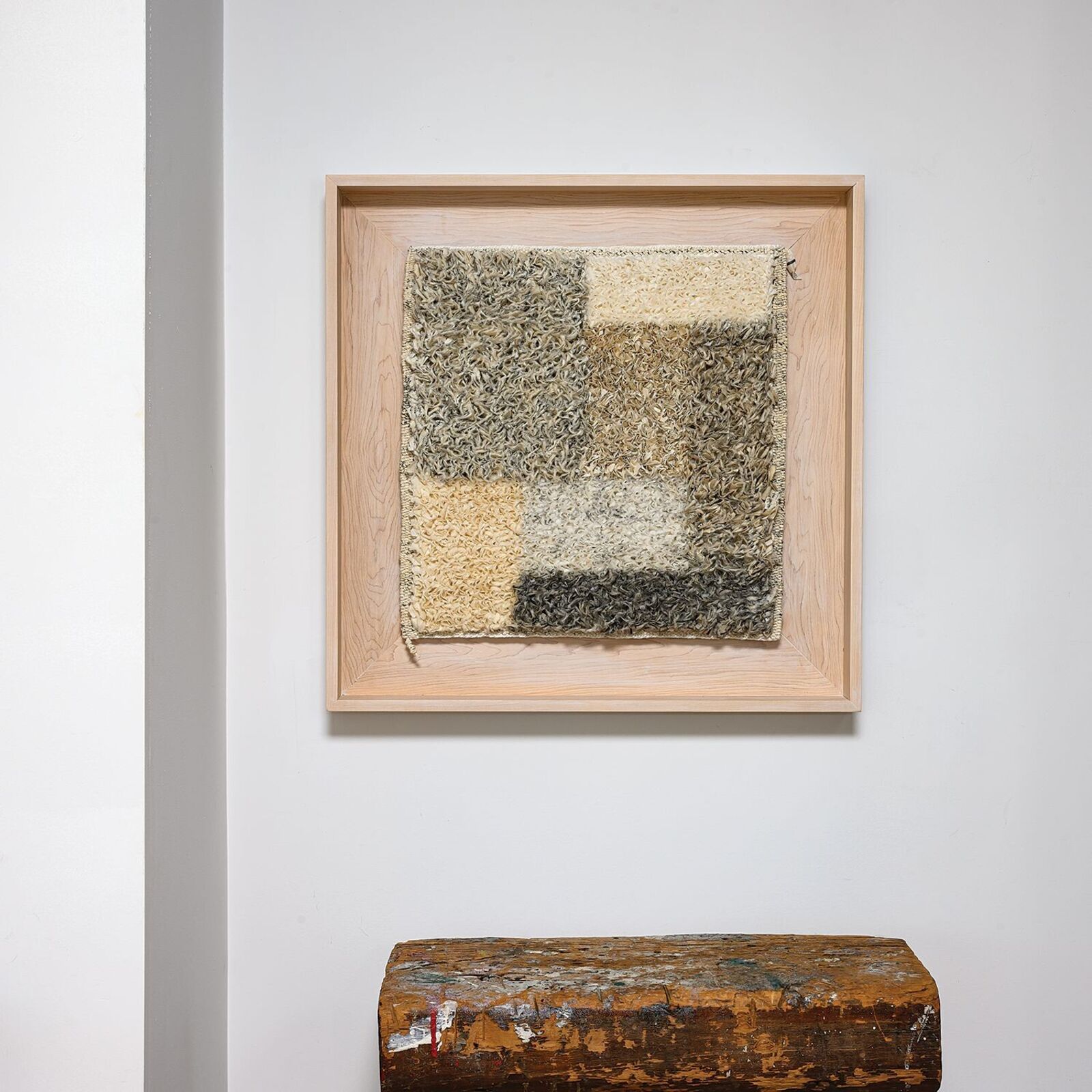 A framed textile artwork featuring a patchwork of textured squares in earthy tones, displayed above a weathered wooden block.