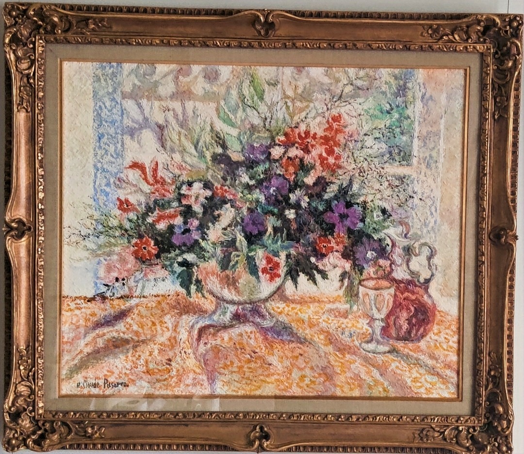 A vibrant still life of a colorful floral arrangement in a bowl, accompanied by a glass and a pitcher on a patterned tablecloth.