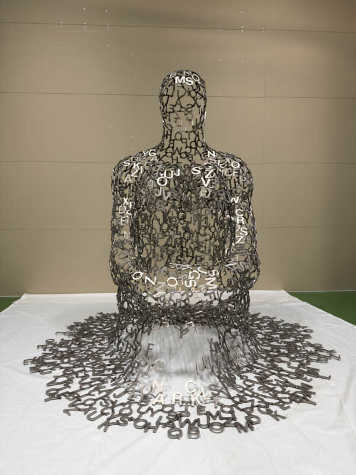 A metallic sculpture of a seated figure constructed from scattered letters, with more letters spilling onto the white floor.