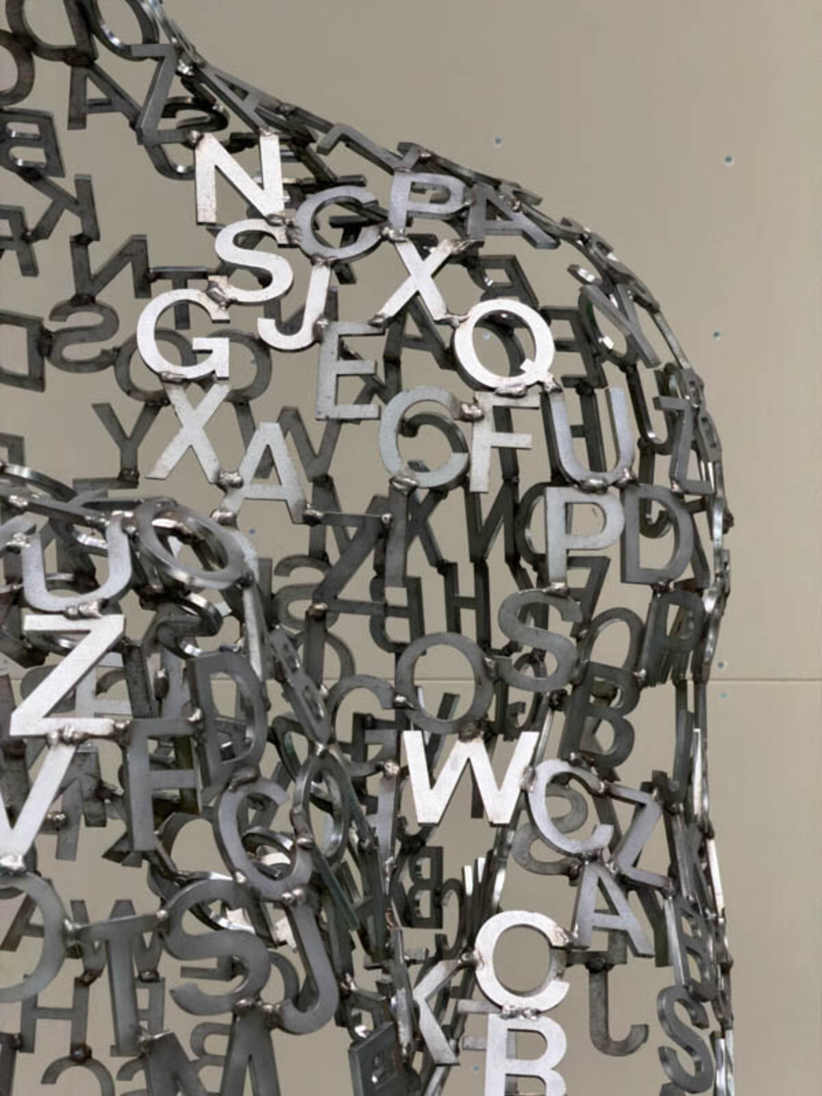 A metallic sculpture of a seated figure constructed from scattered letters, with more letters spilling onto the white floor.