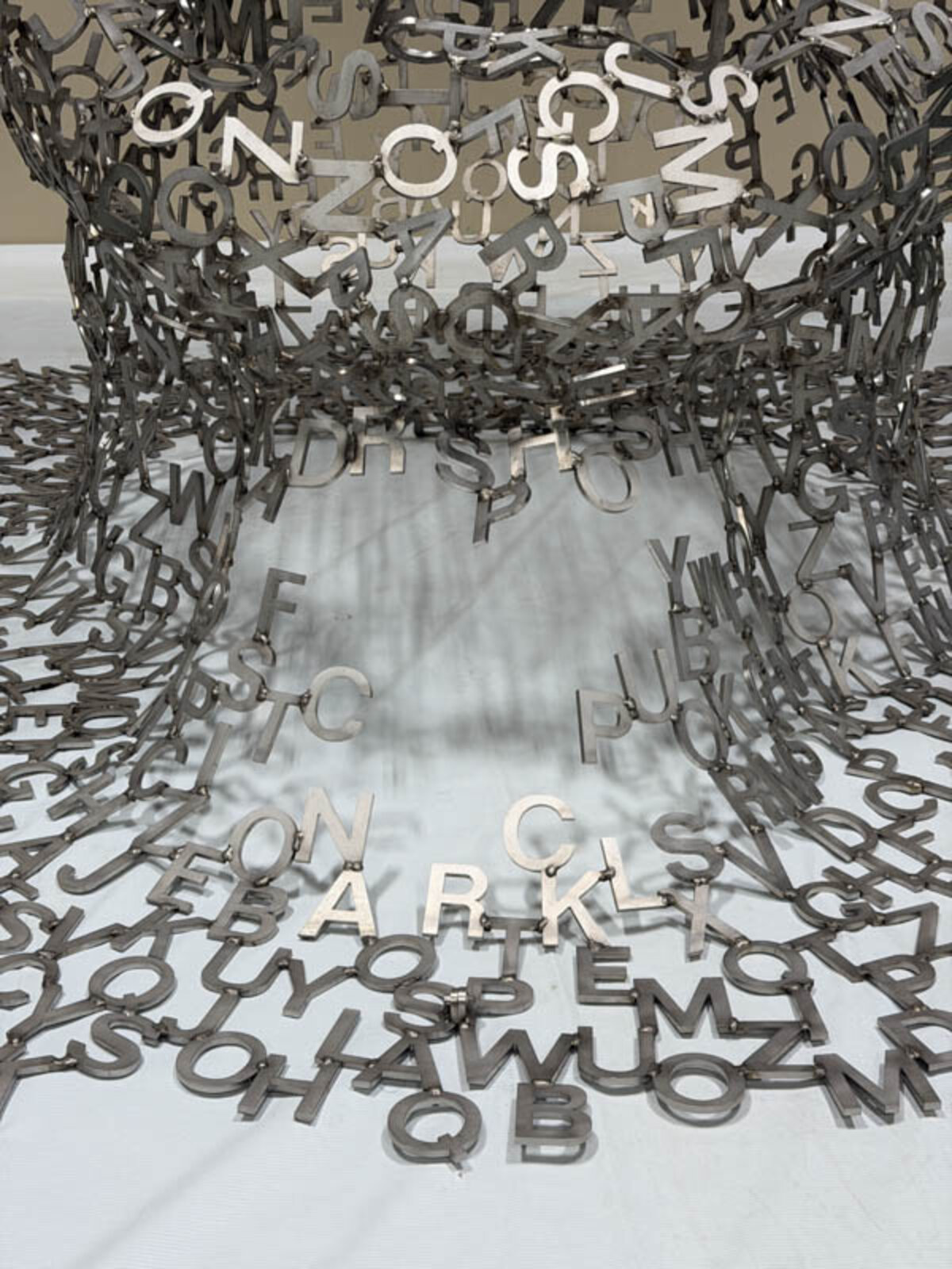 A metallic sculpture of a seated figure constructed from scattered letters, with more letters spilling onto the white floor.