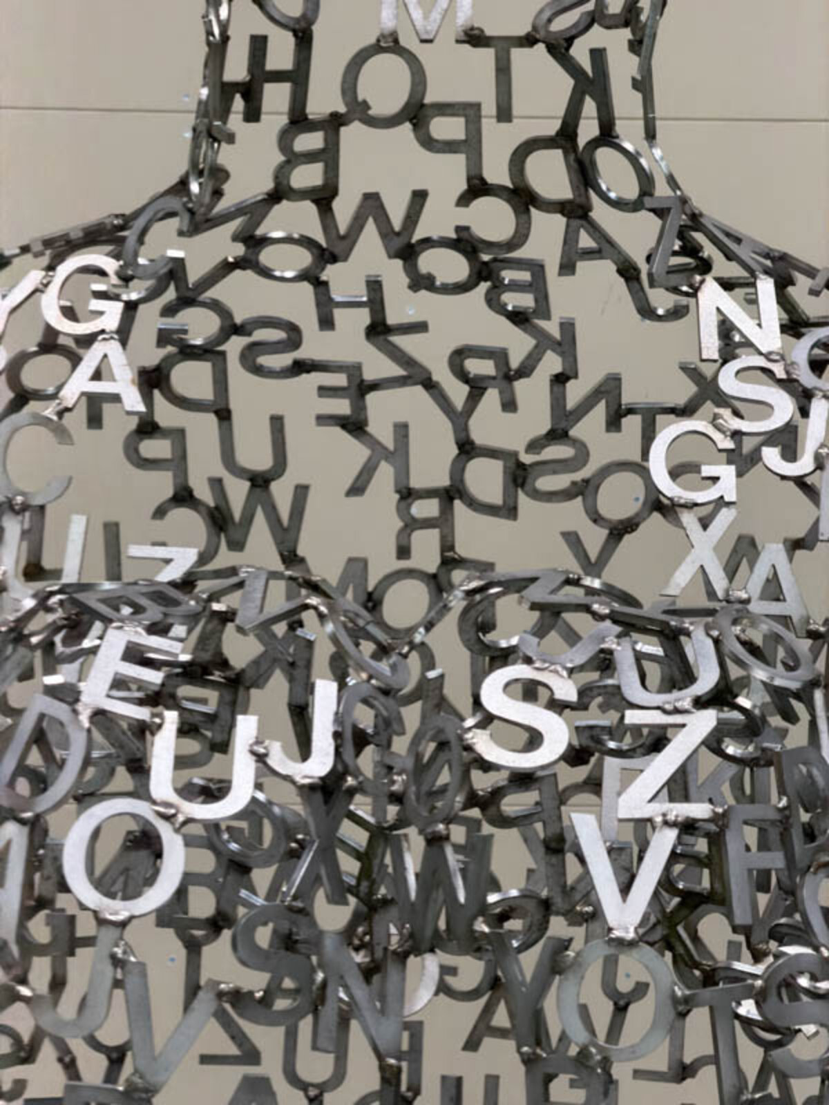 A metallic sculpture of a seated figure constructed from scattered letters, with more letters spilling onto the white floor.