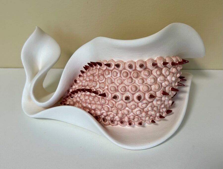 A surreal ceramic sculpture featuring a smooth white vessel containing a fleshy, spiky pink mass.