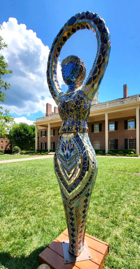 A mosaic sculpture of a figure with an arched halo stands on a wooden base in front of a brick building.