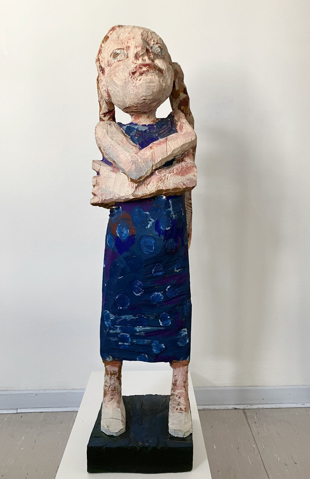 A stylized wooden sculpture of a figure with crossed arms, painted in earthy and deep blue tones.