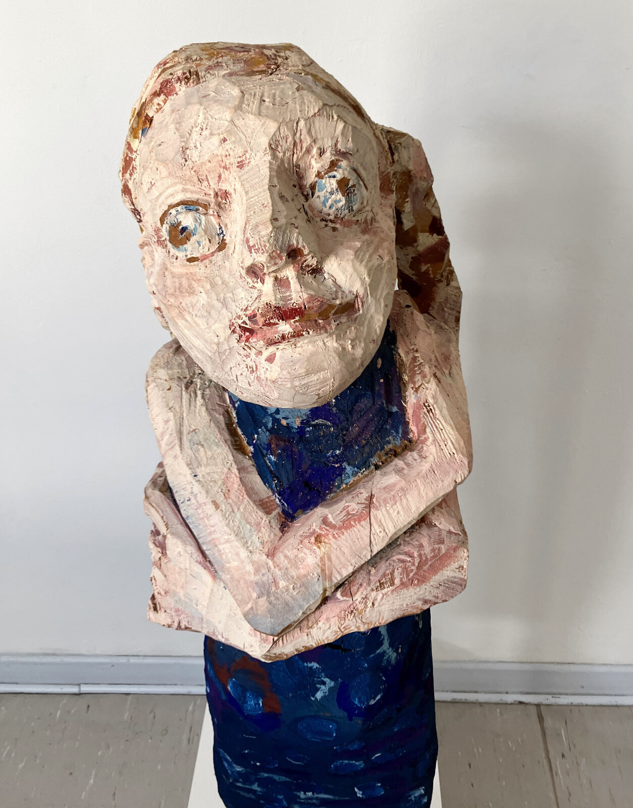A stylized wooden sculpture of a figure with crossed arms, painted in earthy and deep blue tones.