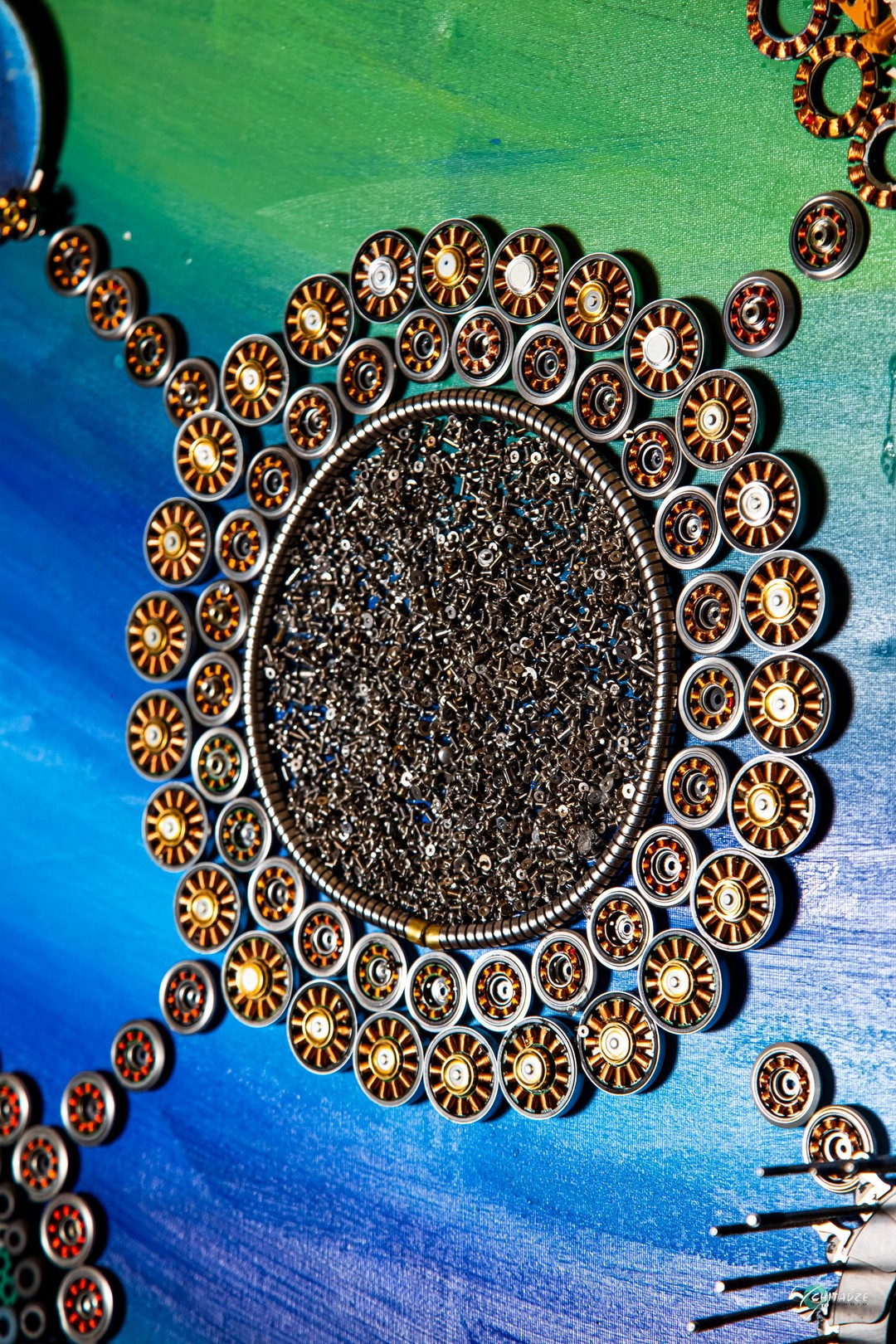 A circular arrangement of metallic discs surrounding a central dark cluster, set against a vibrant blue and green background.