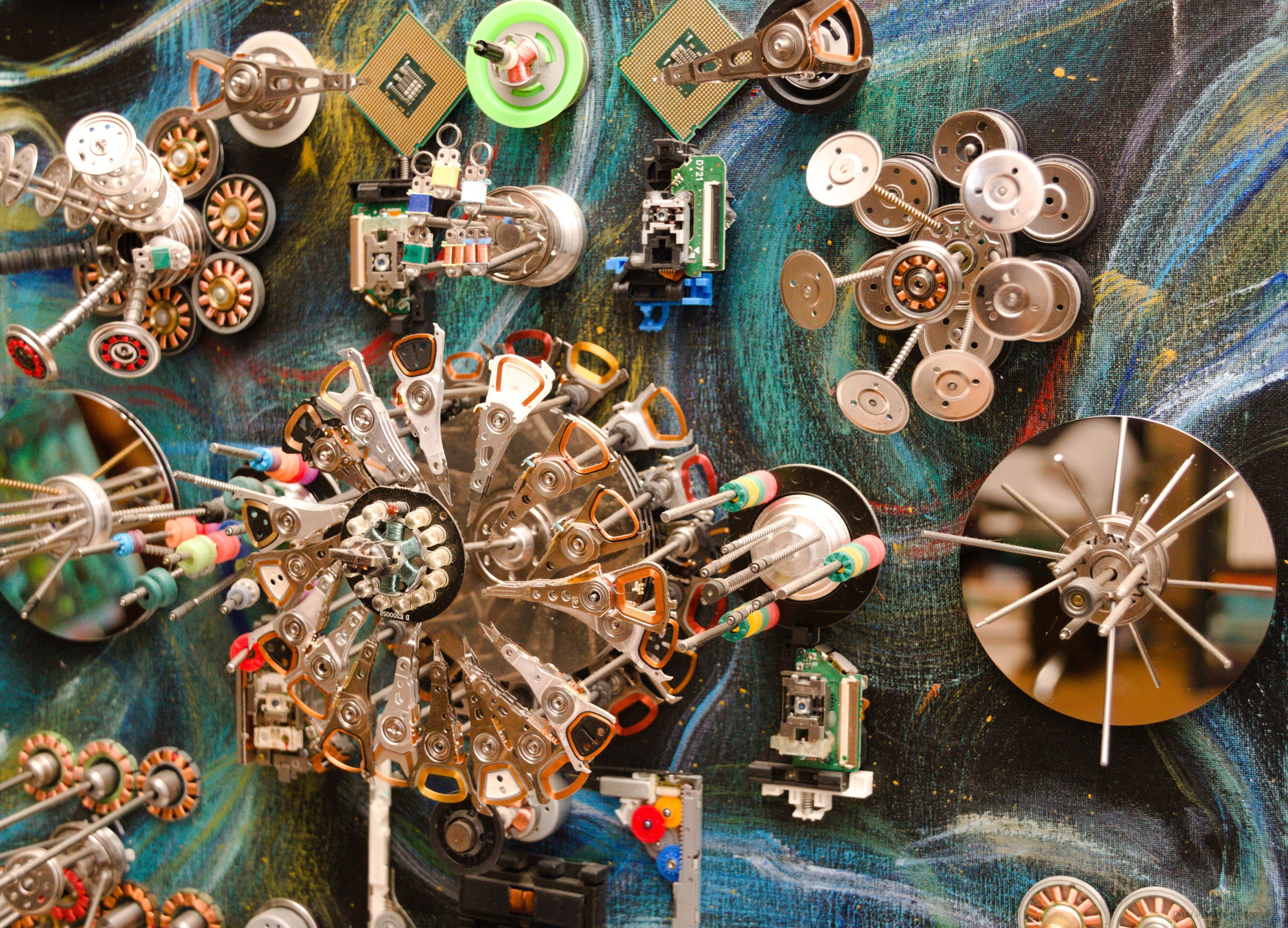 A chaotic collage of metallic mechanical parts and circuit boards scattered across a swirling blue background.
