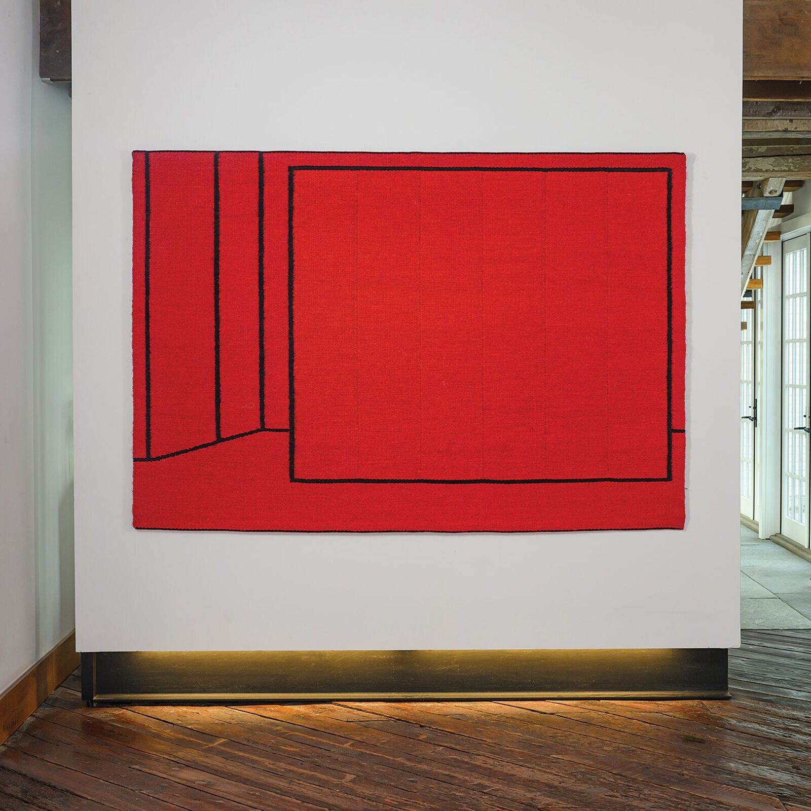 A bold red textile artwork featuring black geometric lines displayed in a gallery space.