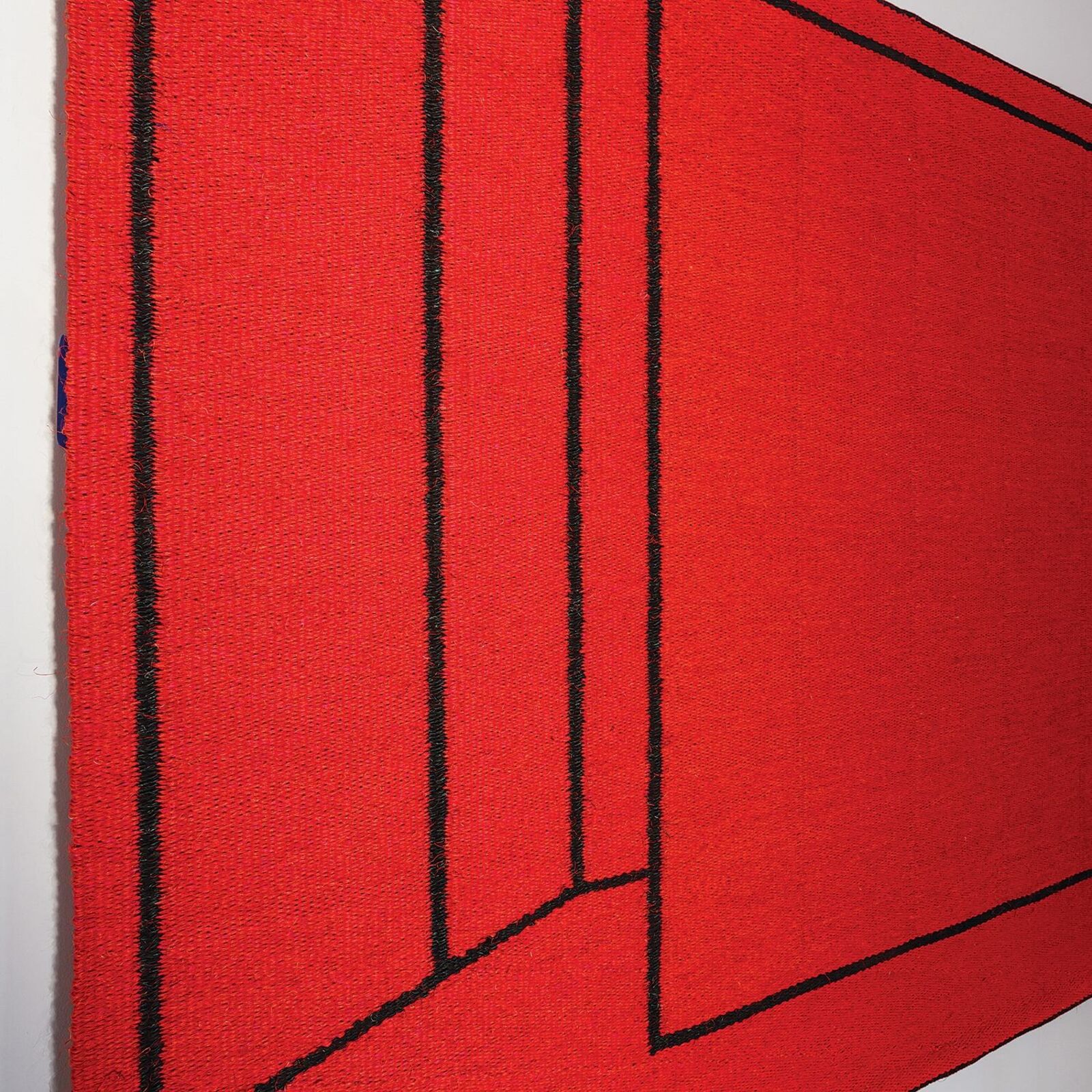 A bold red textile artwork featuring black geometric lines displayed in a gallery space.