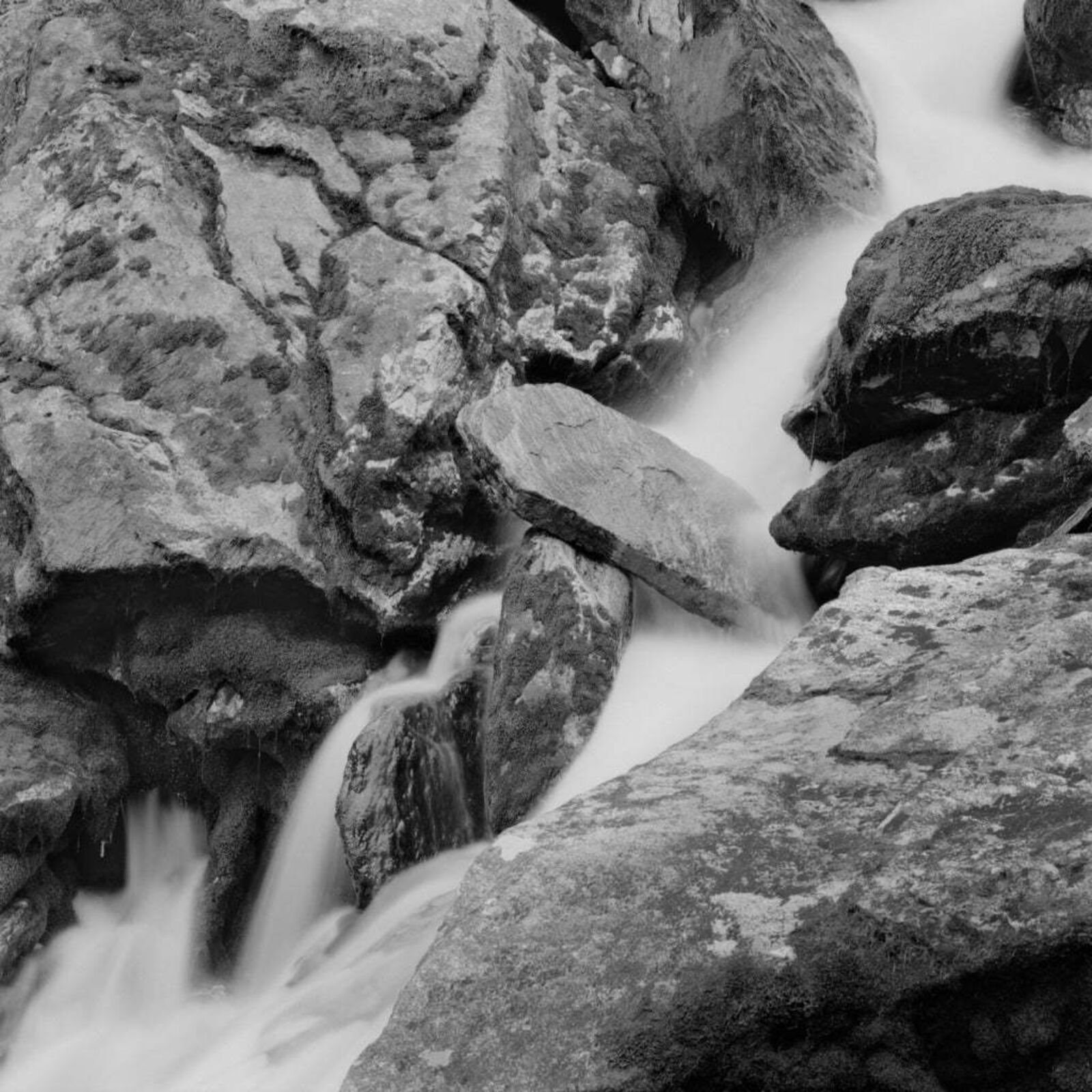 A black and white photograph captures the raw power of a rushing stream flowing through jagged rocks.