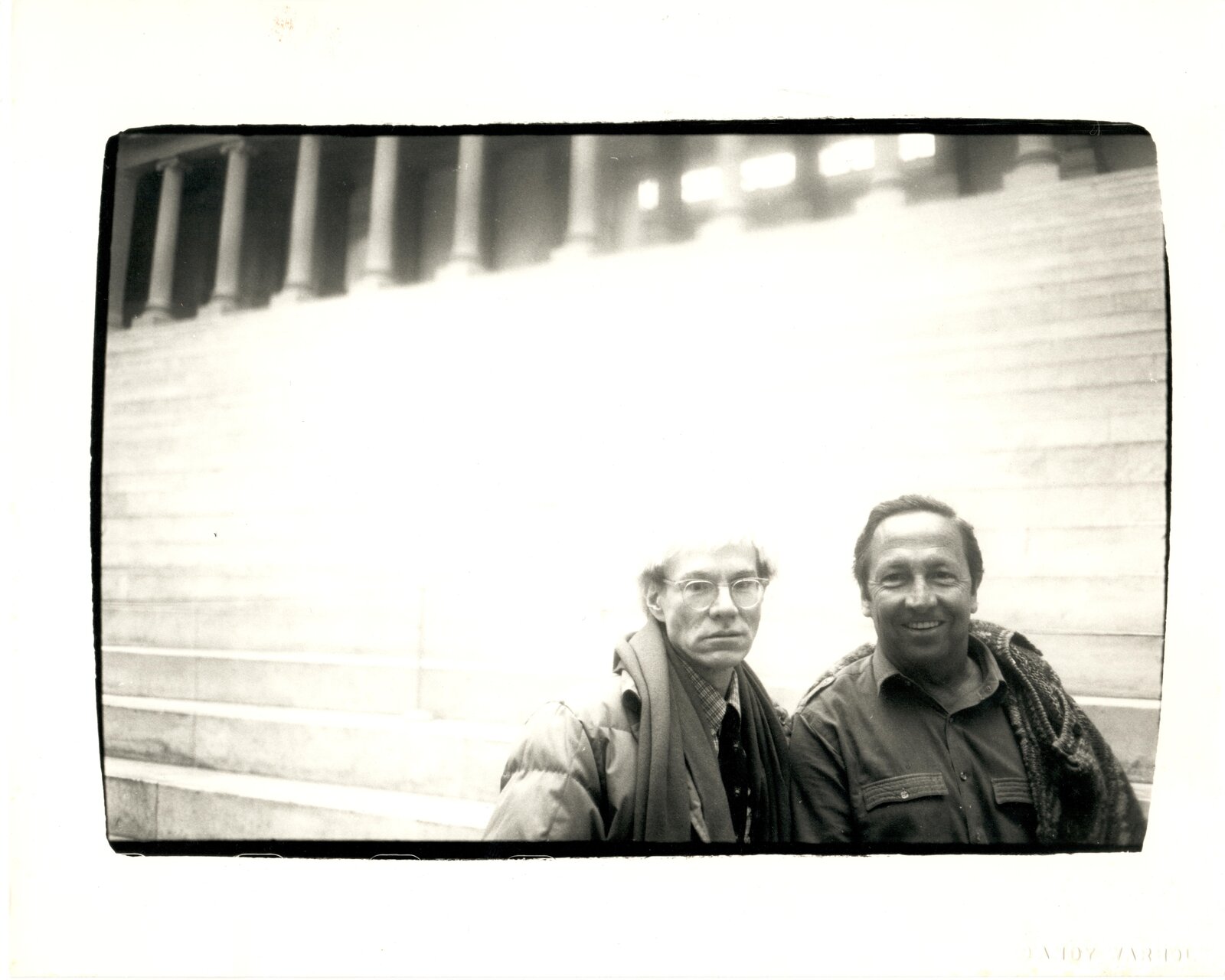 A black-and-white photograph captures two figures standing on wide stone steps before a colonnaded building.