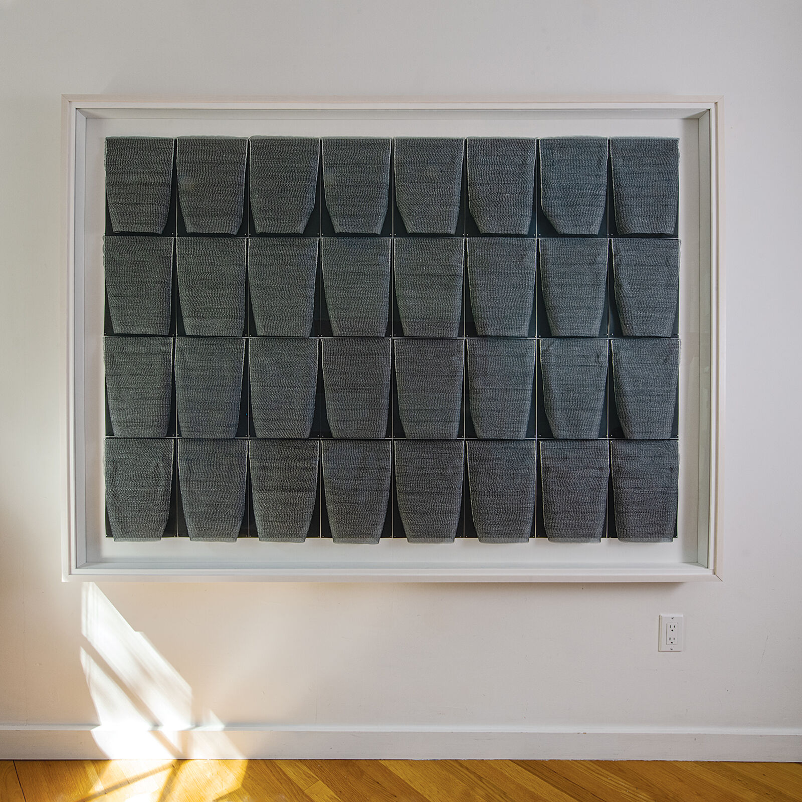 A framed textile grid of grey woven cups on a white wall.