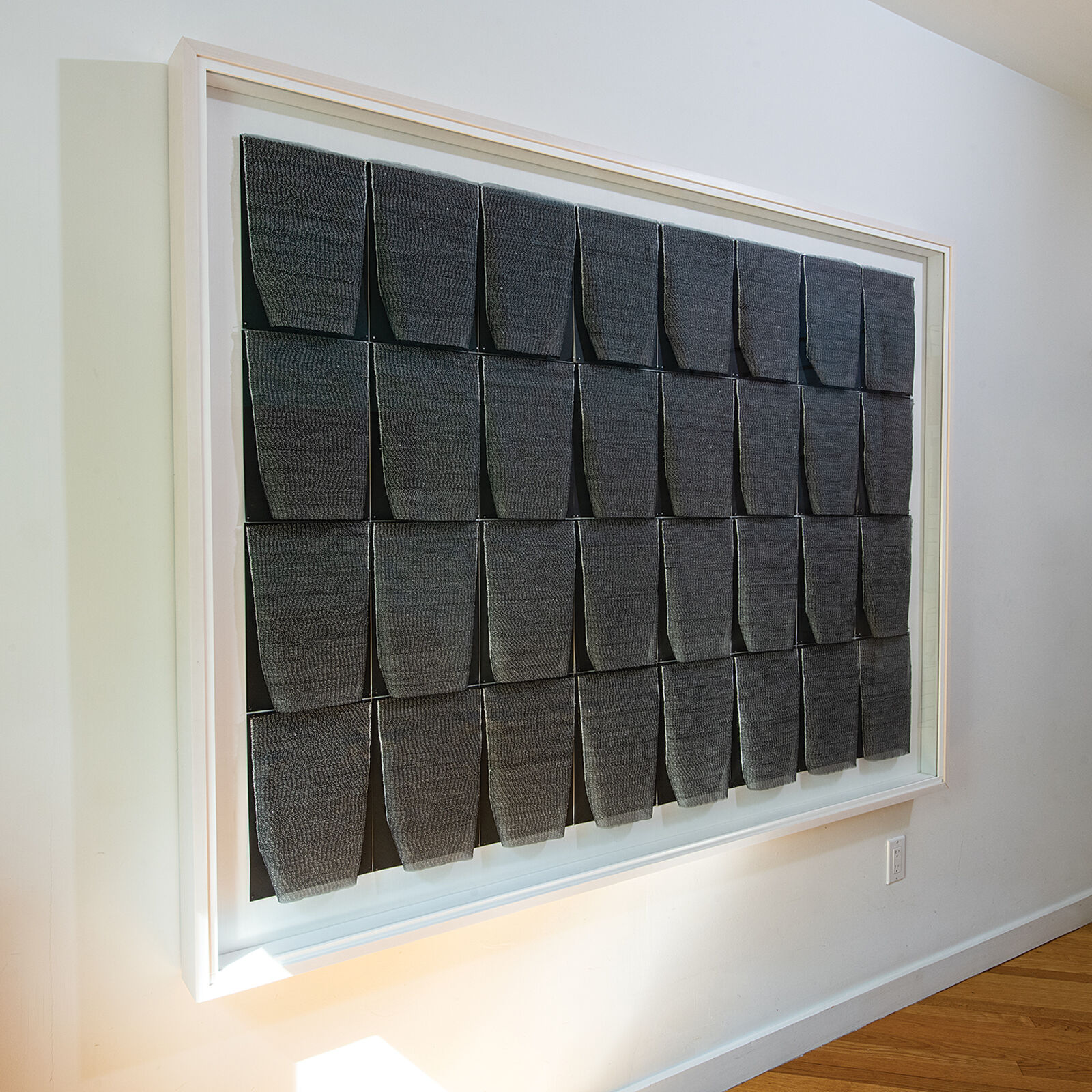 A framed textile grid of grey woven cups on a white wall.