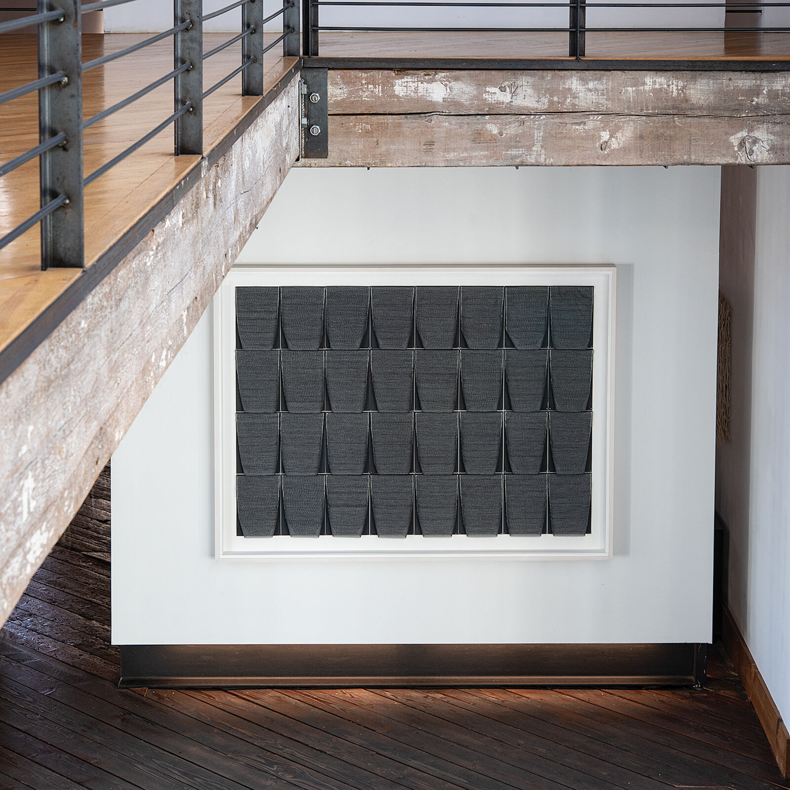 A framed textile grid of grey woven cups on a white wall.