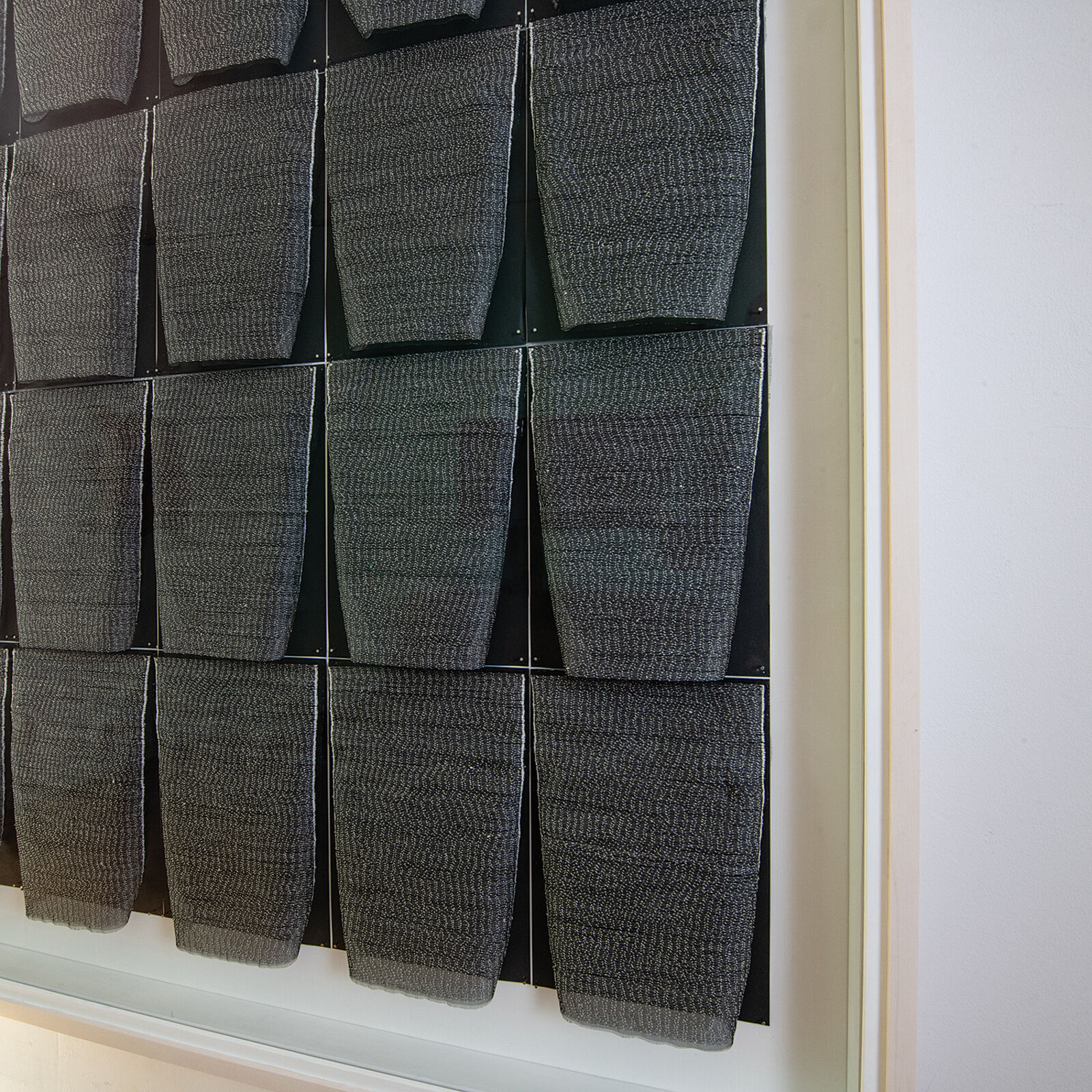 A framed textile grid of grey woven cups on a white wall.