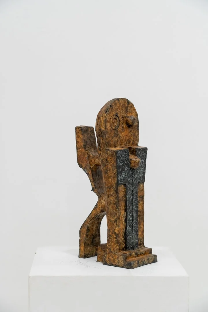 A textured, abstract sculpture of a seated figure with a dark cross-like element on its chest.