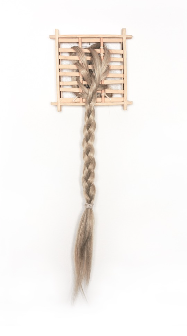 A long braid of light brown hair hangs from a small wooden lattice frame against a stark white background.