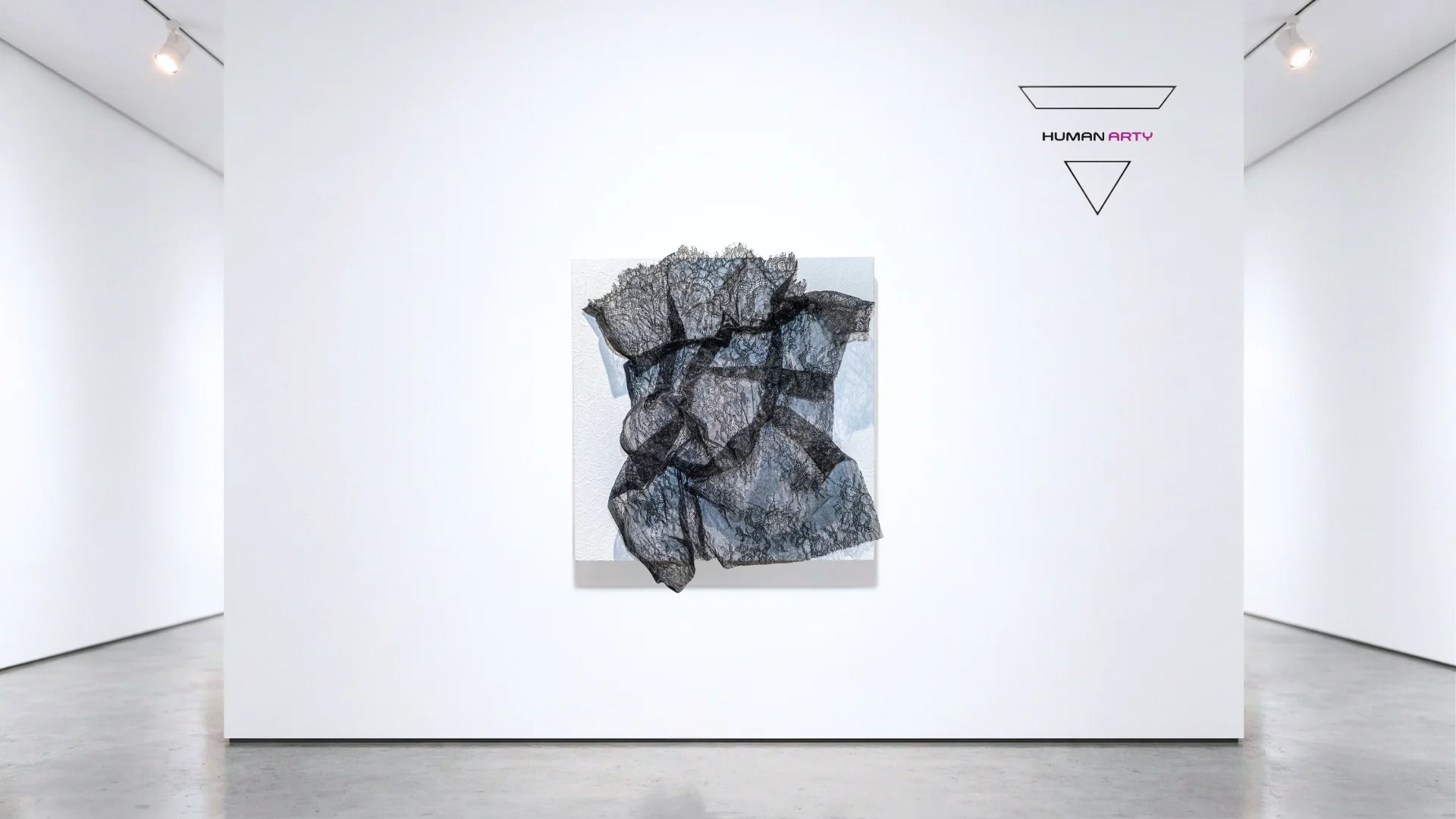 A minimalist gallery scene featuring a black lace sculpture against a stark white wall.
