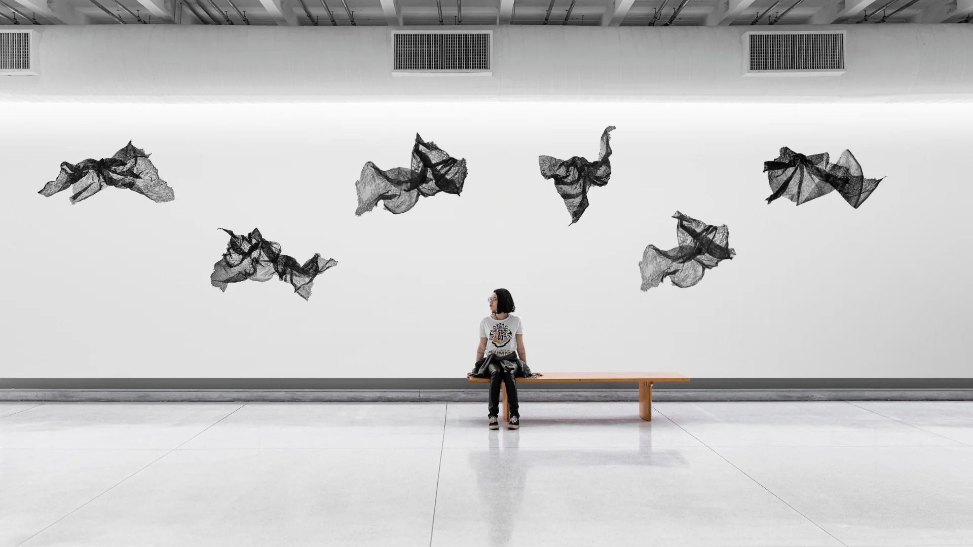 A solitary figure sits on a bench, gazing upward at floating black net-like sculptures against a stark white wall.