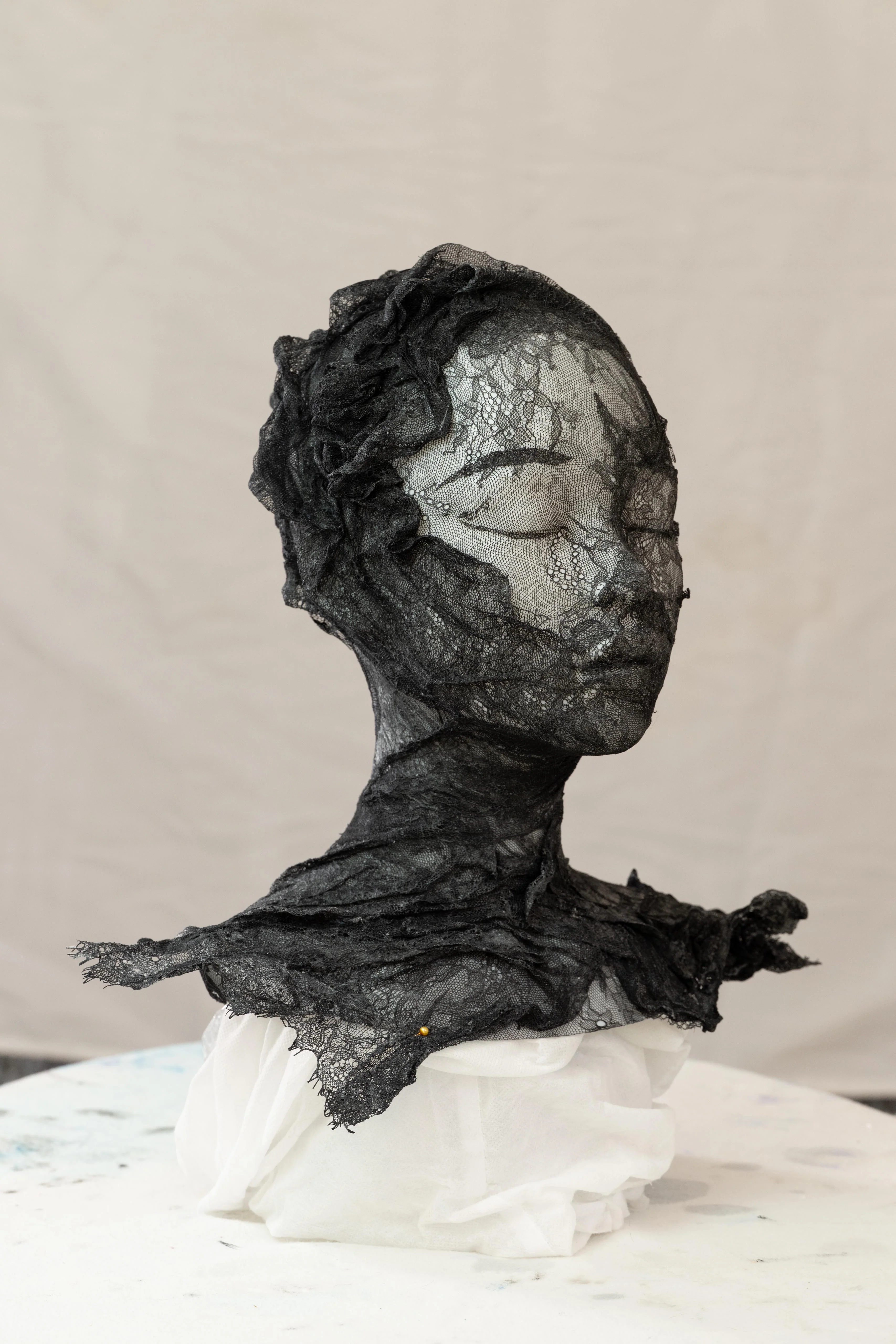 A bust sculpture of a face with closed eyes, crafted from intricate black lace and displayed on a purple pedestal.