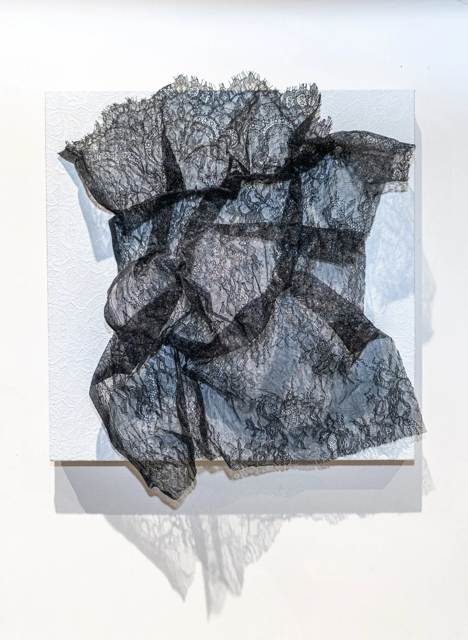 A minimalist gallery scene featuring a black lace sculpture against a stark white wall.