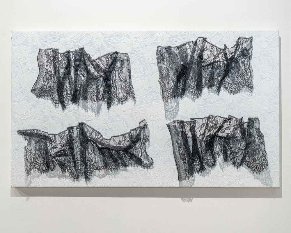 A minimalist gallery scene featuring a monochromatic textile artwork displayed on a white wall.