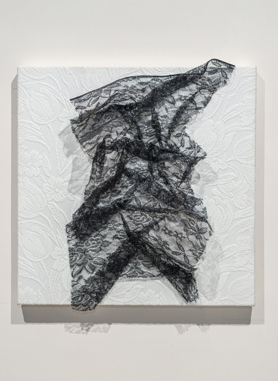 A minimalist gallery scene featuring a square canvas with black lace draped over it.