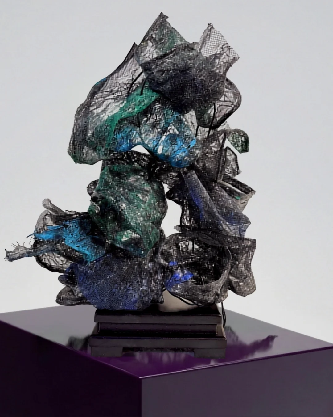 A sculptural lace calligraphy piece with abstract, layered forms in dark tones and hints of blue and green.