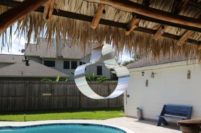 A modern metal sculpture hangs from a thatched roof over a backyard pool.