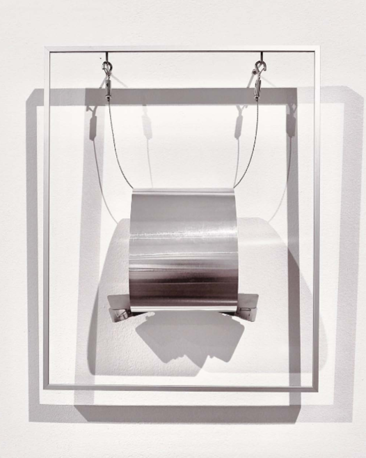 A minimalist sculpture featuring a suspended metallic cylinder within a white frame.