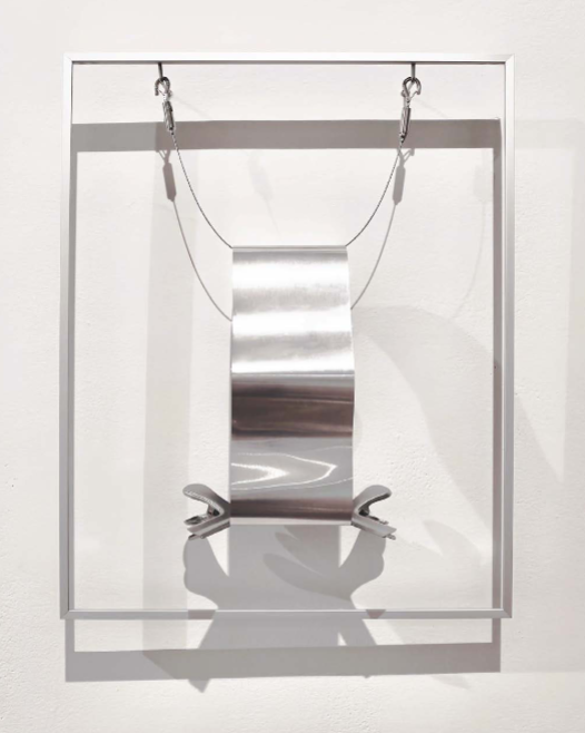 A minimalist sculpture featuring a suspended metallic form within a clear rectangular frame.