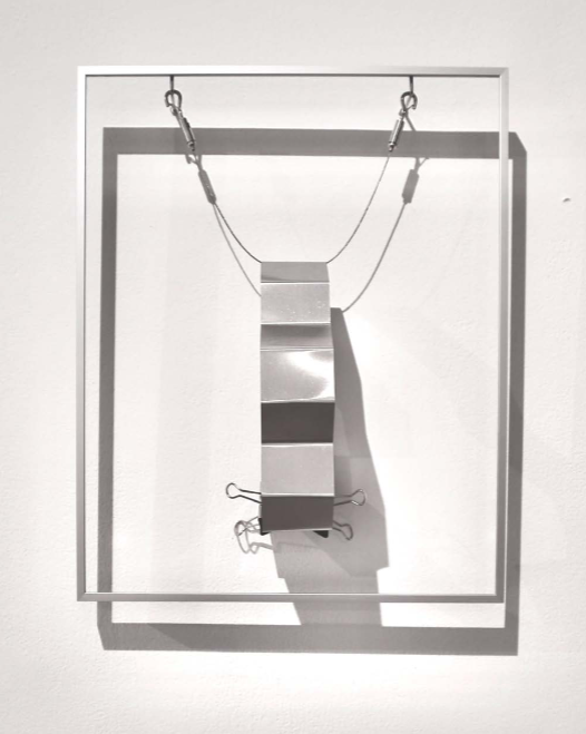 A minimalist sculpture featuring a suspended metallic block within a white frame.