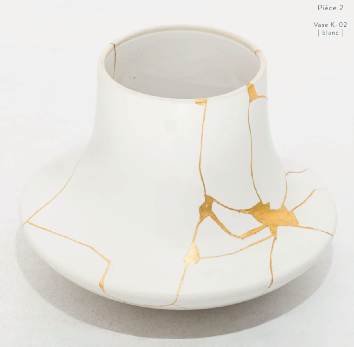 A white ceramic vase with a wide base and narrow neck, featuring a kintsugi-inspired design of gold lines tracing the cracks.